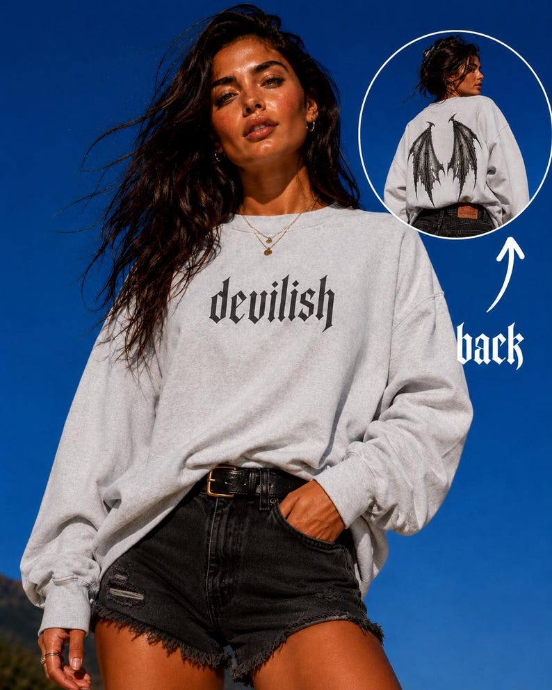 May include: A light gray crewneck sweatshirt featuring the word "devilish" printed in black gothic-style lettering on the front. The back of the sweatshirt displays a large, detailed black graphic of bat-like wings centered between the shoulder blades.