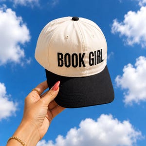 May include: A beige and black baseball cap with the text "BOOK GIRL" in black block letters. The cap is held up against a bright blue sky with white clouds.