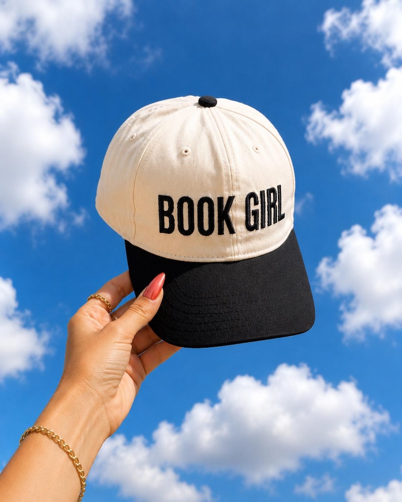 May include: A beige and black baseball cap with the text "BOOK GIRL" in black block letters. The cap is held up against a bright blue sky with white clouds.