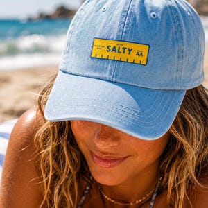 May include: Pale blue denim baseball cap with a curved peak. The cap has a yellow rectangular patch with the word "SALTY" in white and a ruler graphic. The cap is worn on a sunny day at the beach.