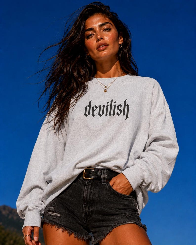 May include: A light grey crewneck sweatshirt featuring the word devilish printed in a black gothic-style font across the chest. The long-sleeved garment is paired with black denim shorts that have a frayed hem and a black leather belt.