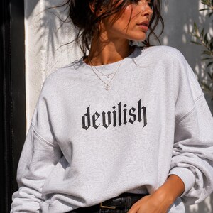 May include: A light grey crewneck sweatshirt featuring the word devilish printed in a black gothic-style font across the chest. The garment has long sleeves with dropped shoulders and a ribbed neckline, cuffs, and hem, providing a relaxed fit.