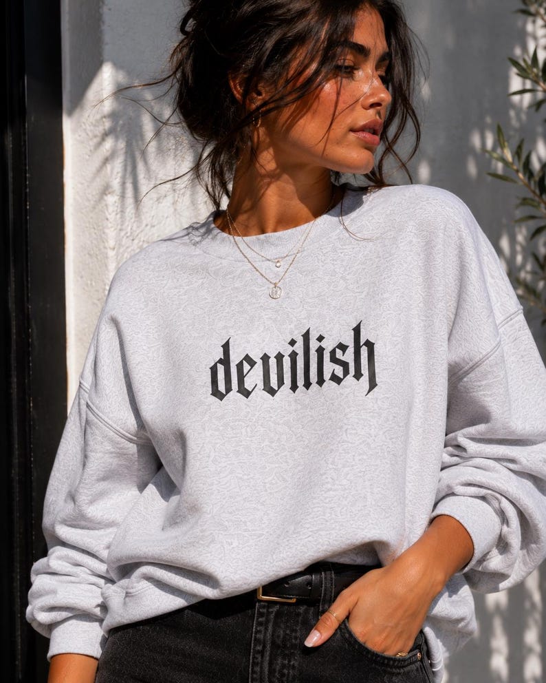 May include: A light grey crewneck sweatshirt featuring the word devilish printed in a black gothic-style font across the chest. The garment has long sleeves with dropped shoulders and a ribbed neckline, cuffs, and hem, providing a relaxed fit.