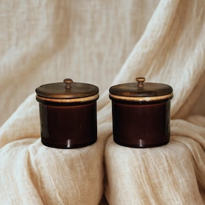 May include: Two cylindrical ceramic jars with a deep brown glossy finish. Each jar features a matching dark brown wooden lid with a small, rounded knob handle at the center. The rims of the jars show a subtle, lighter-toned glazed accent.