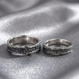 Op de afbeelding: Twee zilveren ringen met een textuur, schorsachtig ontwerp. De ringen staan tegen een zachte, zilvergrijze achtergrond. De ringen zijn waarschijnlijk voor een stel, waarbij de ene iets groter is dan de andere.