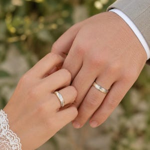 May include: A close-up of two hands, each wearing a wedding ring. The rings are silver with a gold band. The hands are clasped together, suggesting a wedding or engagement. The background is out of focus.