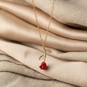 Puede incluir: Un collar dorado con un colgante en forma de rosa roja. La rosa tiene pétalos detallados y está unida a un tallo con dos hojas. La delicada cadena cuelga sobre un fondo de color neutro.