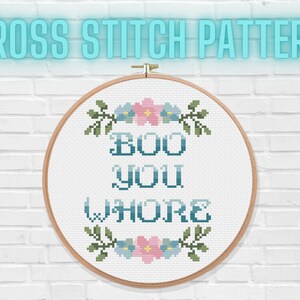 May include: A cross-stitch design in a wooden hoop. The text reads "BOO YOU WHORE" in teal, with floral embellishments. The words "CROSS STITCH PATTERN" are above the hoop in a teal glow, set against a white brick wall.
