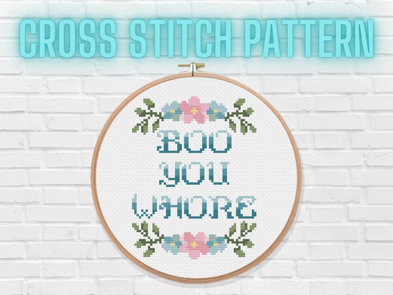 May include: A cross-stitch design in a wooden hoop. The text reads "BOO YOU WHORE" in teal, with floral embellishments. The words "CROSS STITCH PATTERN" are above the hoop in a teal glow, set against a white brick wall.