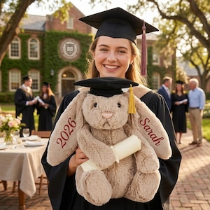 May include: A graduate in a black cap and gown holds a plush bunny wearing a graduation cap. The bunny is light brown with the year "2026" and the name "Sarah" embroidered in burgundy. The bunny holds a rolled-up diploma.