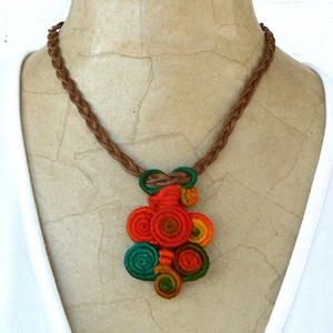 Puede incluir: Un collar hecho a mano y colorido con un cordón trenzado marrón y un colgante con diseños en espiral en rojo, naranja, verde y amarillo. El colgante está unido al cordón con un lazo verde.