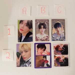 May include: A collection of K-pop idol photo cards. The cards feature images of young men in various poses and outfits. Some cards have decorative borders, while others are plain. The background is a plain white surface.