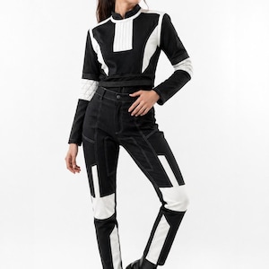 May include: Black and white jumpsuit with geometric paneling. The long-sleeved top features a high collar and white accents. The pants have white panels on the knees and sides. The model is wearing black boots.