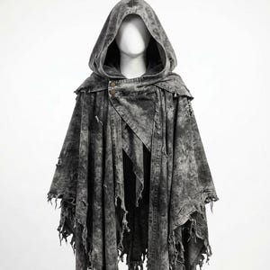 May include: A hooded cape made of distressed grey denim with a washed-out, mottled appearance. The garment features a large hood, two visible buttons at the neckline, and an uneven, frayed hemline with hanging fabric strips throughout the design.