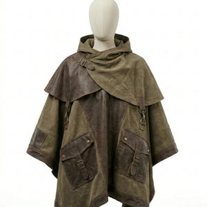 May include: An olive green hooded poncho featuring dark brown leather accents, a wide collar with a buckle strap, and two large front pockets with buckle closures. The garment has a layered design with leather panels and metal hardware details.