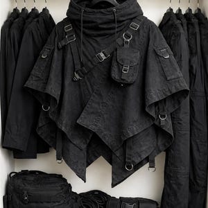 May include: A black hooded poncho featuring an asymmetrical hem, multiple adjustable straps with metal buckles, and a small attached utility pocket on the chest. The garment includes a high collar with drawstring cords and integrated D-ring hardware.