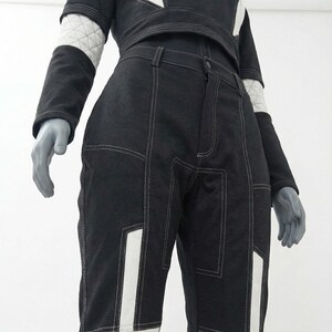May include: Black and white two-piece outfit. The top is a cropped, long-sleeved shirt with white accents on the shoulders and sleeves. The pants are high-waisted with white panels and stitching details. The outfit is displayed on a mannequin.