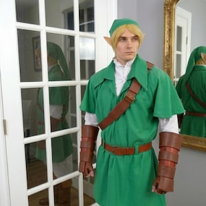 May include: A green tunic with a white undershirt, a green cap, brown leather-like arm guards, a brown belt, and a brown shoulder strap. The outfit includes pointed ear prosthetics and blonde hair, creating a complete fantasy character costume.