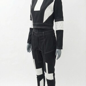 May include: A black and white two-piece outfit on a mannequin. The top is a long-sleeved crop top with white accents and a high collar. The pants are black with white geometric panels and stitching. The outfit is displayed on a gray mannequin.