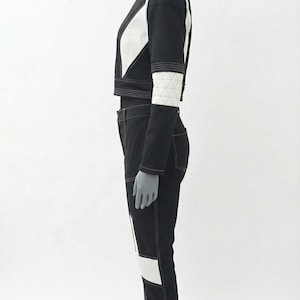 May include: A black and white two-piece outfit on a mannequin. The cropped jacket has white geometric accents and a high collar. The matching pants have white panels and stitching. The outfit is a modern, geometric design.