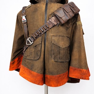 Pu&ograve; includere: Giacca marrone con cappuccio, due tasche frontali e orlo arancione. Presenta una tracolla in pelle con tasche e una fibbia. La giacca &egrave; esposta su un supporto metallico su sfondo bianco.