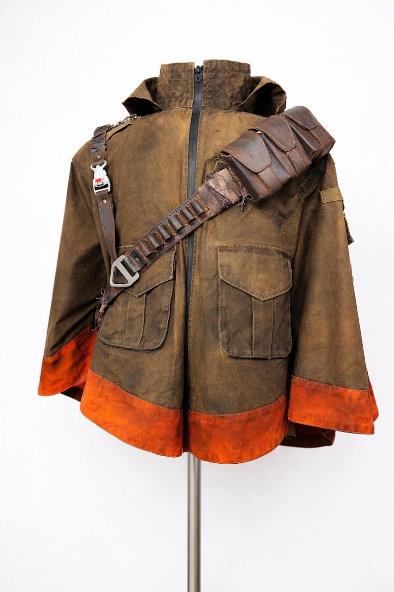 Pu&ograve; includere: Giacca marrone con cappuccio, due tasche frontali e orlo arancione. Presenta una tracolla in pelle con tasche e una fibbia. La giacca &egrave; esposta su un supporto metallico su sfondo bianco.