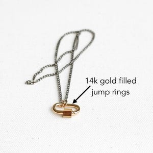 May include: A necklace featuring a dark gray chain attached to a gold-toned oval carabiner clasp with a small square charm. An arrow points to the clasp with the text 14k gold filled jump rings, indicating the material of the connecting components.