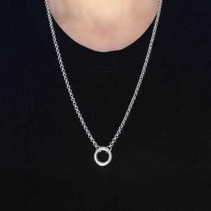 Könnte beinhalten: Eine silberne Halskette mit einem kleinen, kreisförmigen Anhänger. Die Kette besteht aus kleinen, runden Gliedern. Die Halskette wird vor einem schwarzen Hintergrund präsentiert.