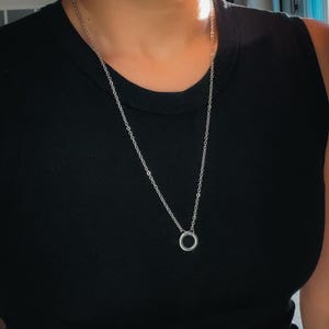 May include: A silver-colored necklace with a small, circular pendant. The necklace is worn over a black sleeveless top. The chain is delicate and the pendant is a simple, minimalist design.