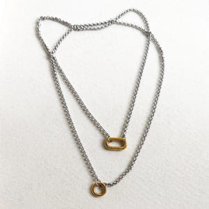 Puede incluir: Un collar de dos capas con dos cadenas. La cadena superior presenta un colgante ovalado dorado, mientras que la cadena inferior tiene un colgante circular dorado. Las cadenas son plateadas y tienen un diseño delicado y enlazado.