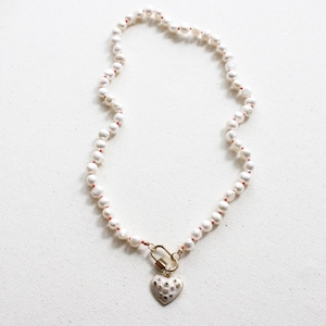 May include: A pearl necklace with a gold heart pendant. The necklace features white pearls strung on a red thread. The heart pendant is gold with small, embedded crystals. The necklace is displayed on a white surface.