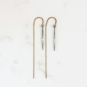 May include: A pair of delicate threader earrings. The earrings feature a gold-toned curved hook and a silver-colored, elongated, cylindrical charm. The chain is gold-toned. The earrings are displayed on a white marble surface.