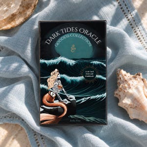 May include: A rectangular Dark Tides Oracle Goddess Collection card deck box featuring an illustration of a skeleton mermaid sitting on a rock amidst dark teal ocean waves under a moonlit sky. The box includes a 46 card deck and guidebook.