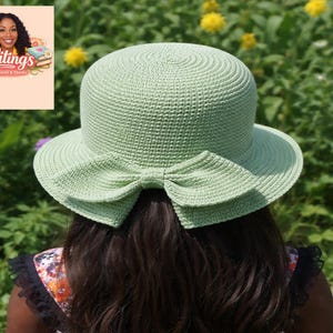 Puede incluir: Un sombrero de ganchillo verde claro con un lazo en la parte trasera. El sombrero tiene un ala ancha y una copa redonda. La persona que lleva el sombrero tiene el pelo oscuro. El fondo es un jardín con flores amarillas.