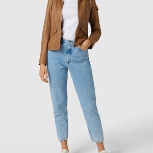 May include: A woman wearing a brown leather jacket with two front pockets, a white t-shirt, light blue jeans, and white sneakers. The jacket has a collared neckline and a zipper closure. The jeans are cuffed at the ankles.
