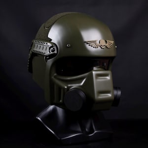 May include: An olive green military-style helmet with a matte finish. The helmet features a visor, a respirator, and a gold-coloured emblem of a skull with wings. The helmet is displayed on a black stand against a black background.