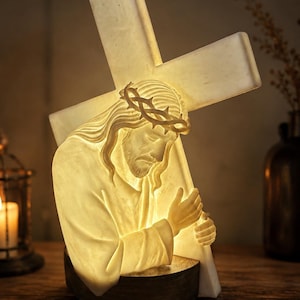 May include: A glowing, off-white sculptural lamp depicting a figure with long hair wearing a crown of thorns, leaning forward while carrying a large cross. The translucent material emits a warm light, highlighting the detailed facial features.