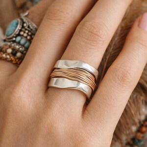 May include: A silver ring with a wavy design, wrapped with multiple strands of gold-colored wire. The ring is on a hand, with other rings visible in the background. The ring is a statement piece of jewelry.
