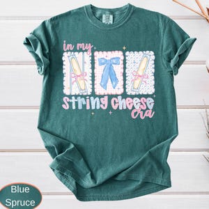 May include: Blue spruce t-shirt with a graphic design. The design features the text "in my string cheese era" in pink and white, along with three illustrated rectangles. Two rectangles have string cheese illustrations, and one has a blue bow.