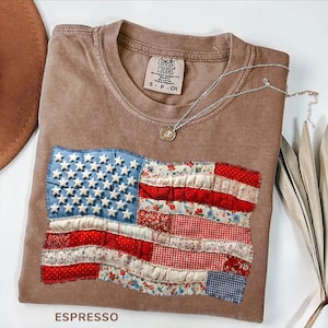 Op de afbeelding: Espresso-kleurig T-shirt met een opgenaaid Amerikaans vlagontwerp. De vlag heeft blauwe stof met witte sterren en rood-wit gestreepte stof. Een zilveren ketting met een hanger ligt op het shirt. Het shirt is gelabeld "Comfort Colors."