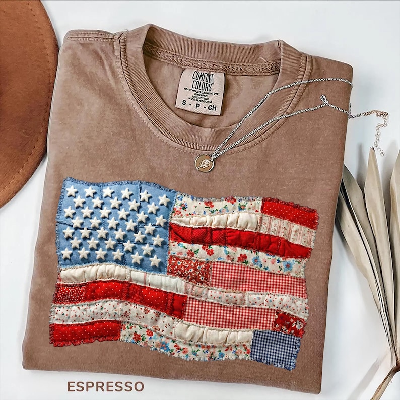 Op de afbeelding: Espresso-kleurig T-shirt met een opgenaaid Amerikaans vlagontwerp. De vlag heeft blauwe stof met witte sterren en rood-wit gestreepte stof. Een zilveren ketting met een hanger ligt op het shirt. Het shirt is gelabeld "Comfort Colors."