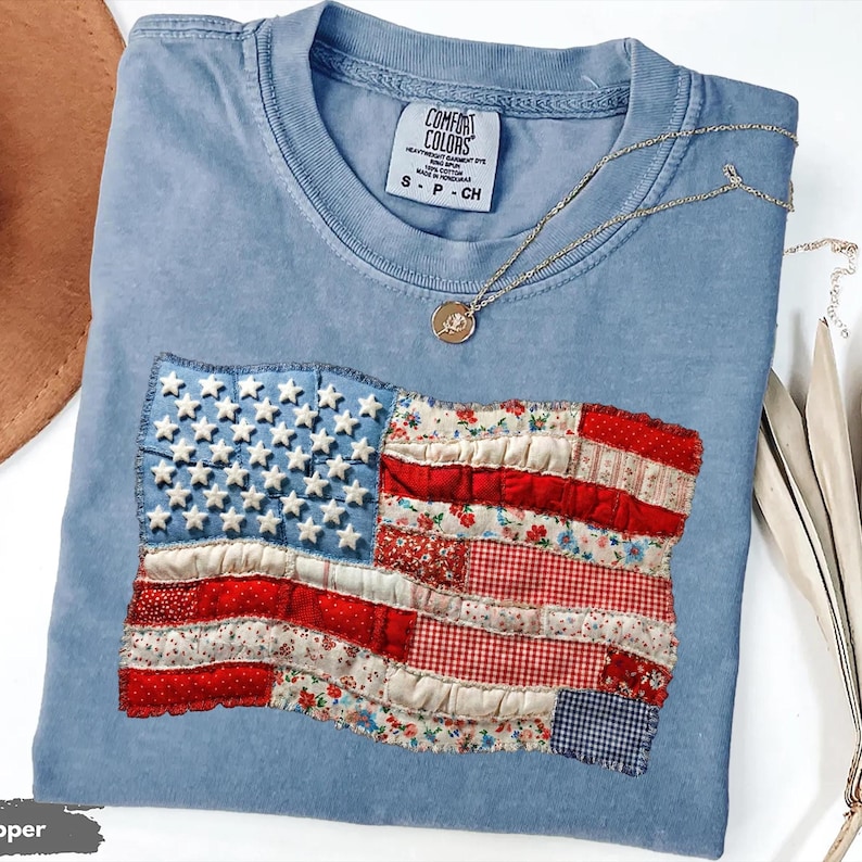 An illustration mimicking a handmade quilt styled as the American flag. It displays stitched panels of floral, gingham, and dotted fabrics forming the stripes. Plump textured stars sit on a frayed woven background.