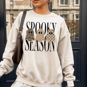 May include: A cream-colored crewneck sweatshirt featuring the black text SPOOKY SEASON. Three ghost-shaped patches are centered on the chest, each with a different pattern: one in green camouflage, one in tan leopard print, and one in a tan checkerboard.