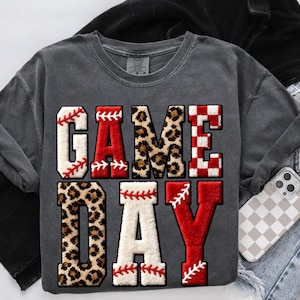 May include: A charcoal gray short-sleeve t-shirt featuring the text GAME DAY in large, textured, varsity-style letters. The letters incorporate various patterns including white baseball stitching, solid red, brown leopard print, and a red-and-white checkerboard.