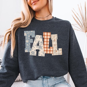 May include: A dark charcoal gray crewneck sweatshirt featuring the word FALL in large block letters across the chest. Each letter has a distinct pattern: the F is blue floral, the A is cream floral, the L is orange gingham, and the L is plaid.