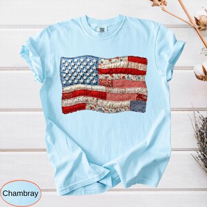 Op de afbeelding: Lichtblauw T-shirt met een patchwork ontwerp van de Amerikaanse vlag. De vlag heeft een blauw denim veld met witte sterren, rood-wit gestreepte secties en bloemenstof. Het woord "Chambray" staat in een cirkel linksonder.