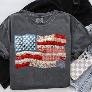 Op de afbeelding: Een donkergrijs sweatshirt met een patchwork Amerikaans vlagontwerp. De vlag bevat denim, bloemen en gingham stoffen voor de sterren en strepen. Het sweatshirt wordt getoond met een spijkerbroek en een telefoonhoesje met een dambordpatroon.