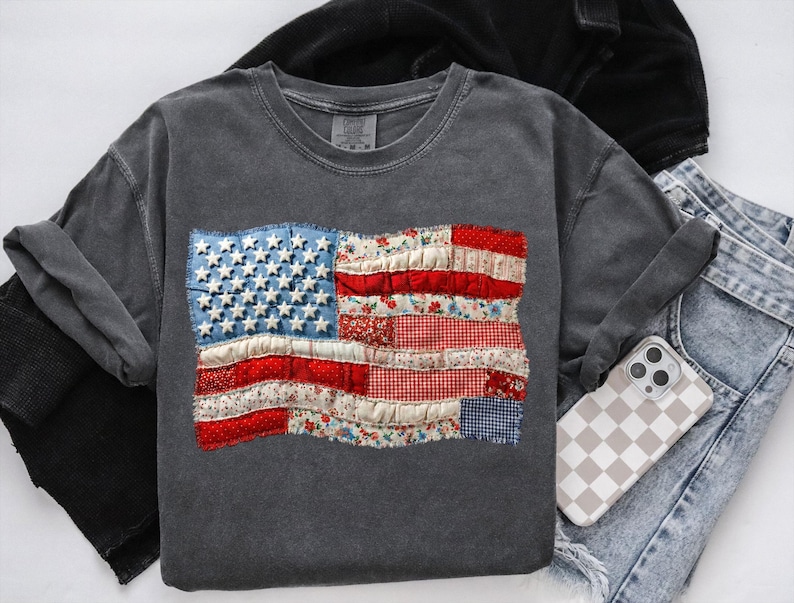 Op de afbeelding: Een donkergrijs sweatshirt met een patchwork Amerikaans vlagontwerp. De vlag bevat denim, bloemen en gingham stoffen voor de sterren en strepen. Het sweatshirt wordt getoond met een spijkerbroek en een telefoonhoesje met een dambordpatroon.
