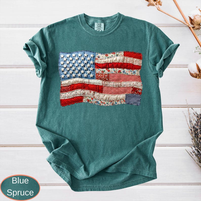 Op de afbeelding: Een tealkleurig T-shirt met een patchwork ontwerp van de Amerikaanse vlag. De vlag gebruikt stoffen met rode, witte en blauwe patronen, wat een distressed look geeft. Het shirt heeft een ronde hals en korte mouwen. De kleur is Blue Spruce.