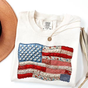 Op de afbeelding: Cr&egrave;mekleurig T-shirt met een patchwork ontwerp van de Amerikaanse vlag. De vlag bevat blauwe denim met witte sterren en rood- en bloempatroon strepen. Een gouden ketting en een bruine hoed zijn ook zichtbaar.